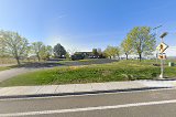 Low-resolution Google Street View-style exterior shot of a low-rise commercial building at a road intersection surrounded by trees.